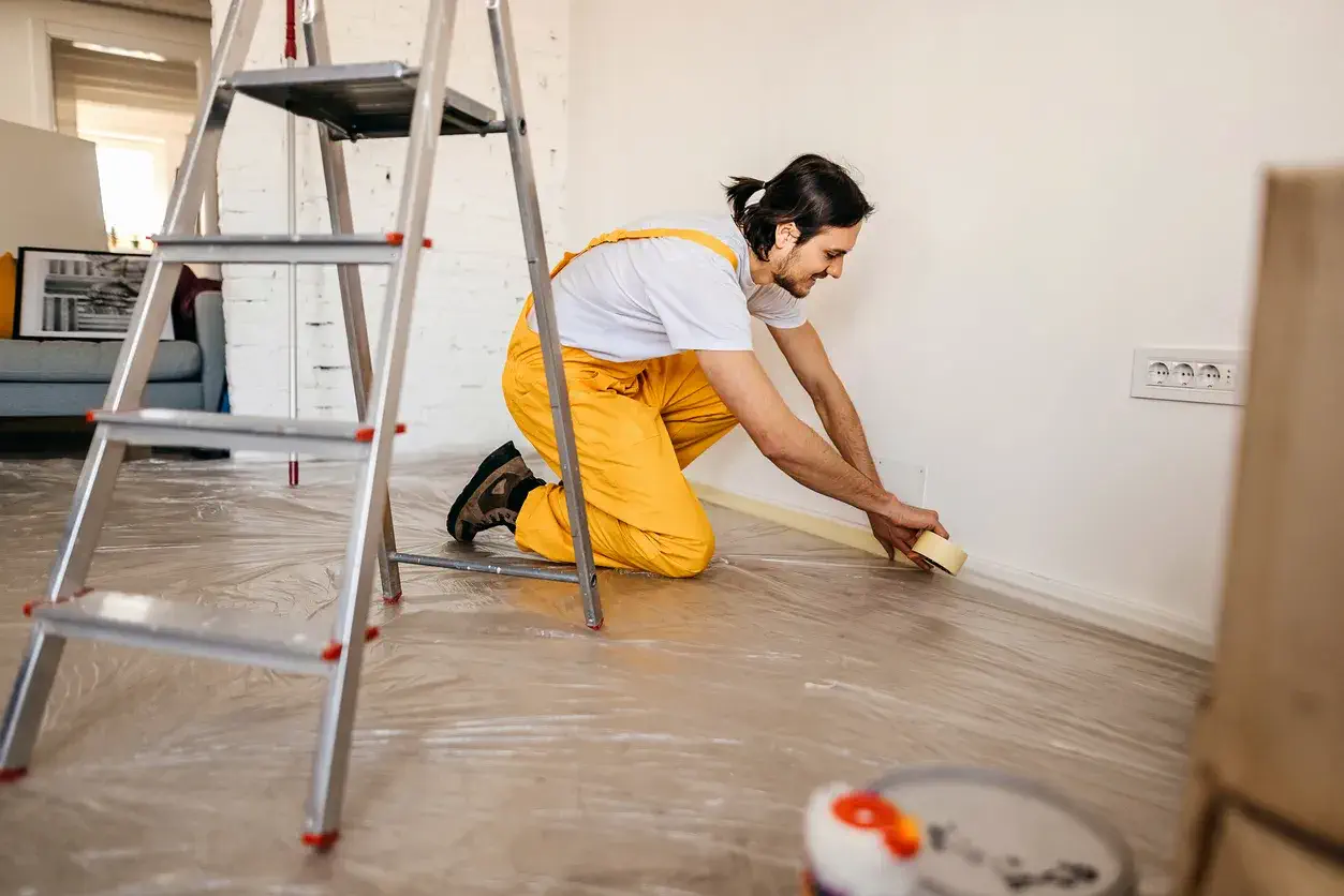 Boya badana işlemi öncesinde süpürgeliklere maskeleme bandı uygulayarak boya hazırlığı yapan sarı tulumlu profesyonel usta